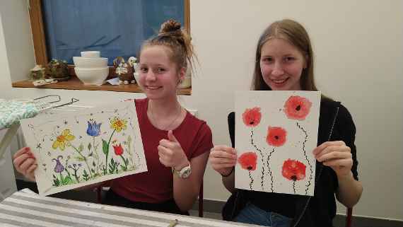 Zwei Schülerinnen halten ihre gemalten Bilder hoch und lächeln 