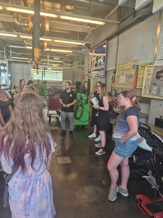Werkstattbesichtigung: Ein Mann mit schwarzem T-Shirt erklärt einer Gruppe von Frauen eine Motorradwerkstatt