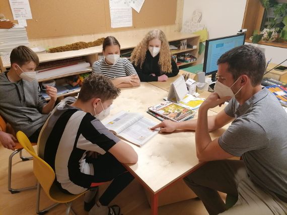 Schülerinnen und Schüler sitzen mit einem Lehrer beim Tisch und lernen 